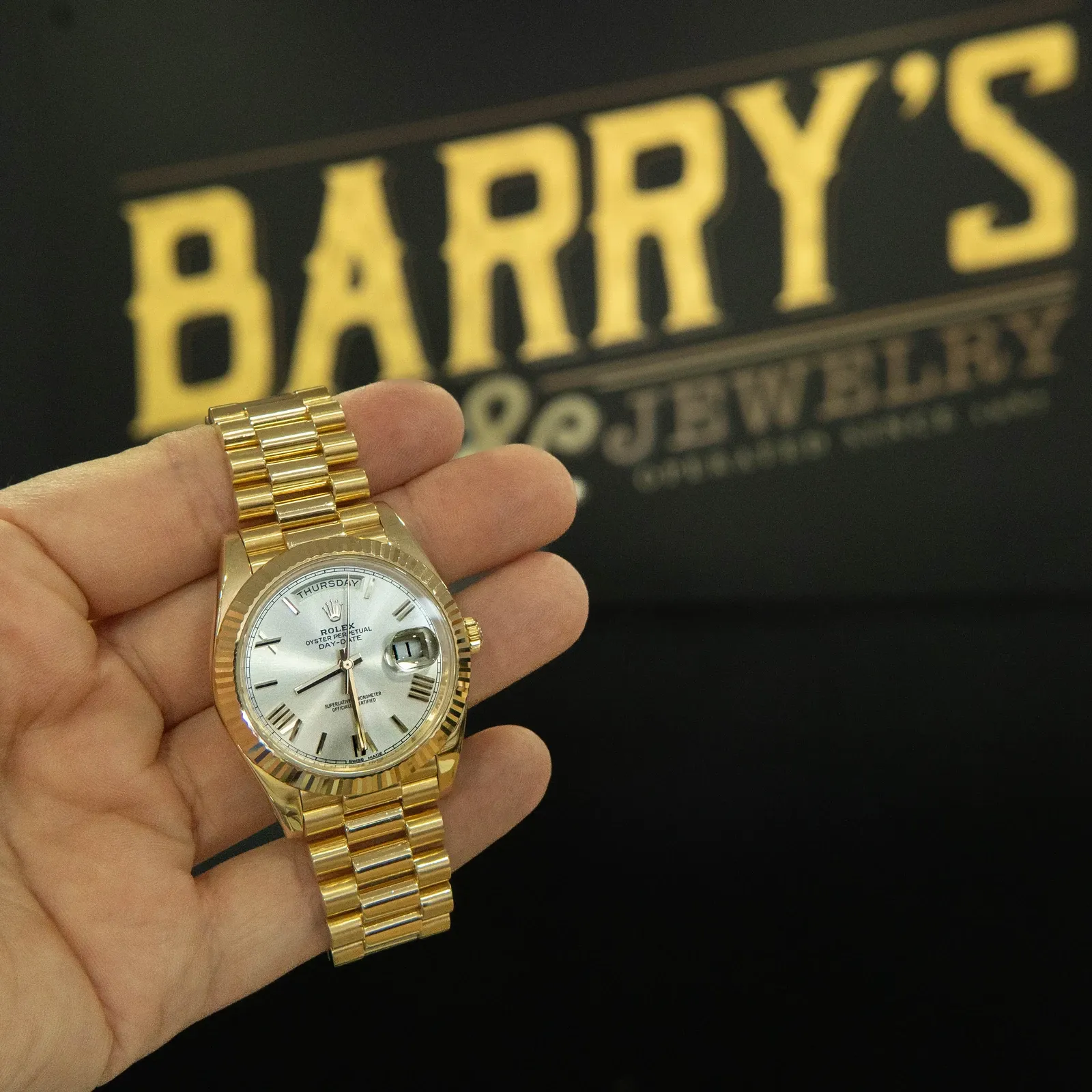A gold Rolex Day-Date held up in front of the Barry's Pawn & Jewelry sign inside the Boca Raton store
