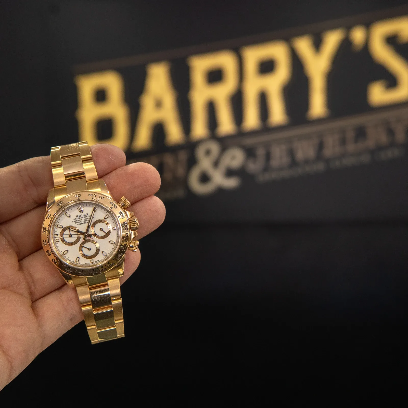 A gold Rolex Daytona with a white dial held up in front of the Barry's Pawn & Jewelry sign inside the Boca Raton store