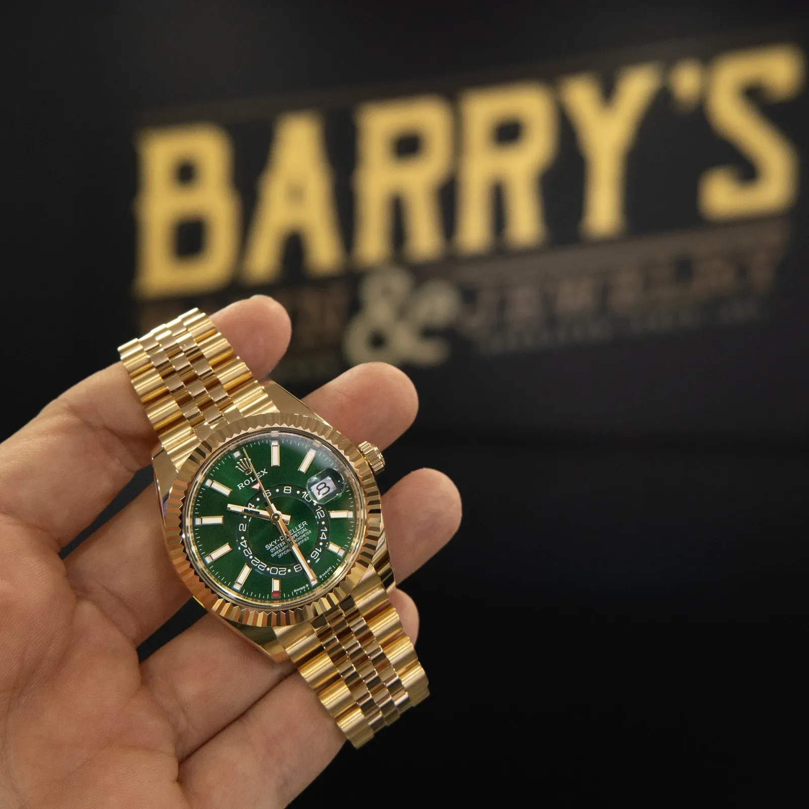 A gold Rolex Sky-Dweller with a green dial held up in front of the Barry's Pawn & Jewelry sign inside the Boca Raton store