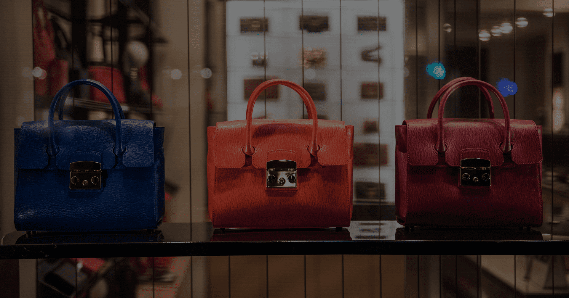 Three handbags in blue, orange, and burgundy are displayed on a shelf in a store.