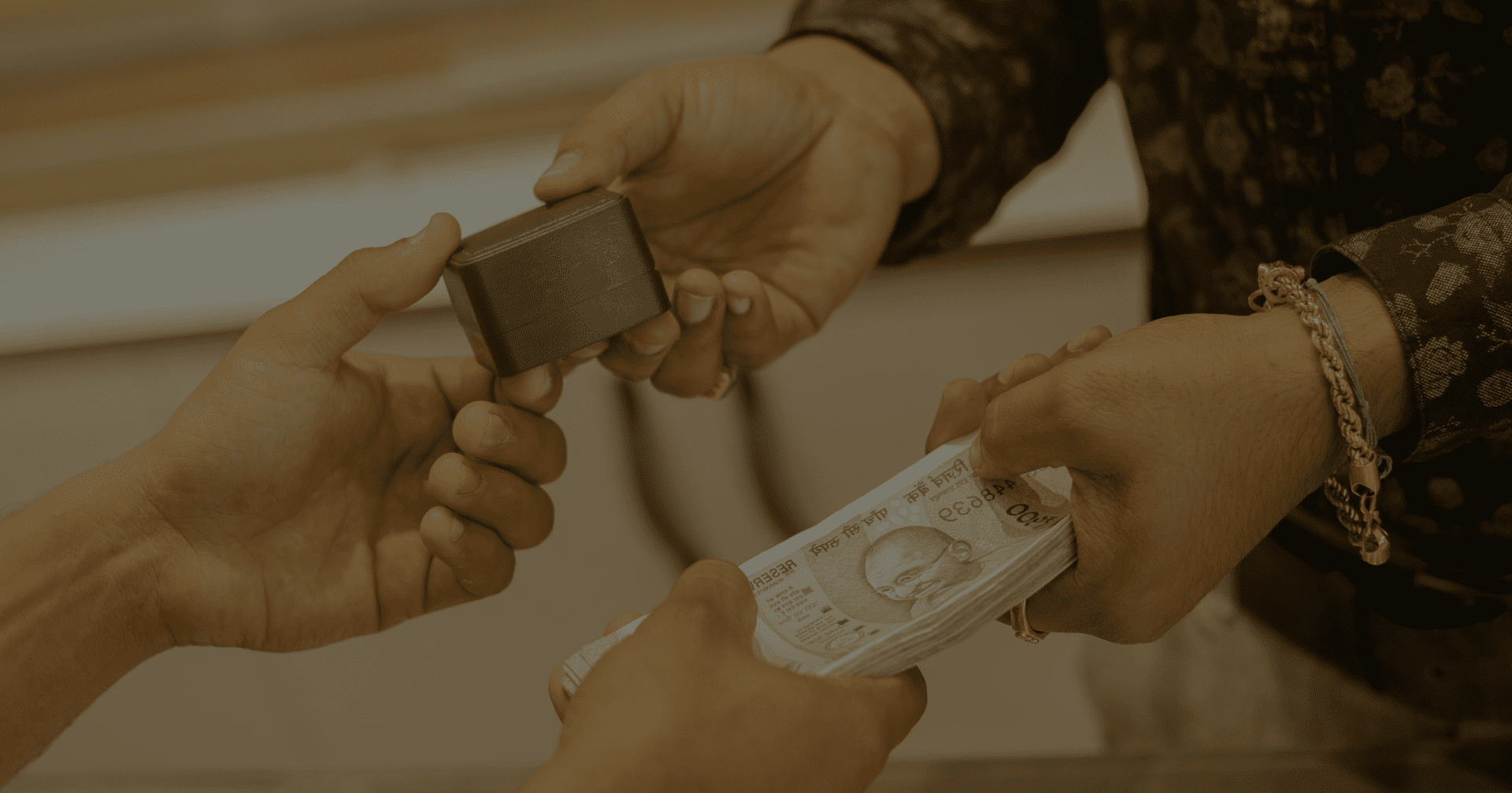 Two hands exchange a stack of banknotes for a small rectangular object. One person wears a patterned shirt and bracelet, while the other two hands are bare.