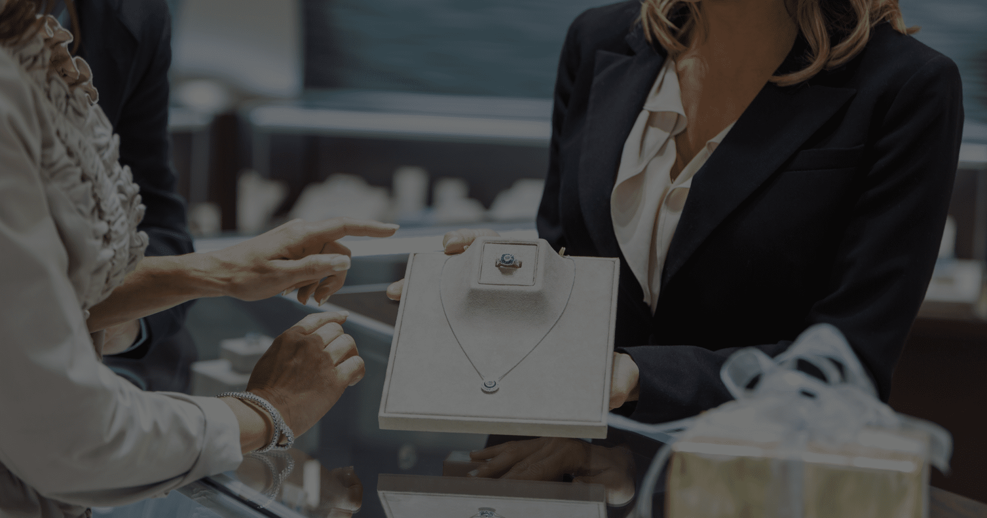 A person holds a necklace set on a display board in a jewelry store, while another person points to it over the counter.