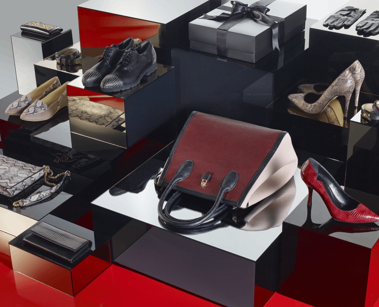 Display of luxury fashion accessories including various shoes, a handbag, gift boxes, and a wallet on reflective surfaces.