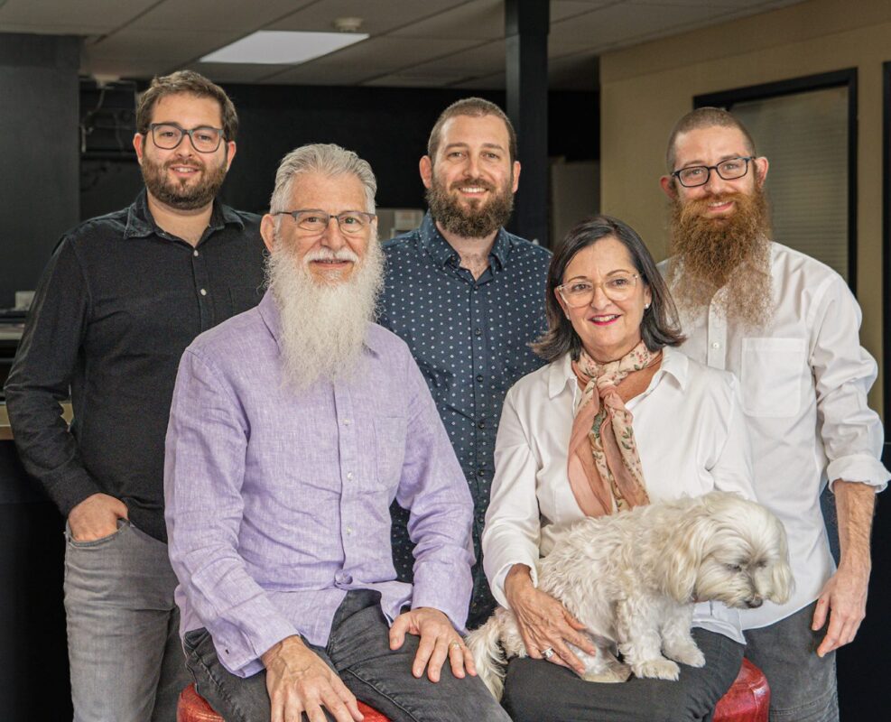Group of five adults and a small white dog posing indoors. The individuals are smiling, and one older man and one woman are seated.