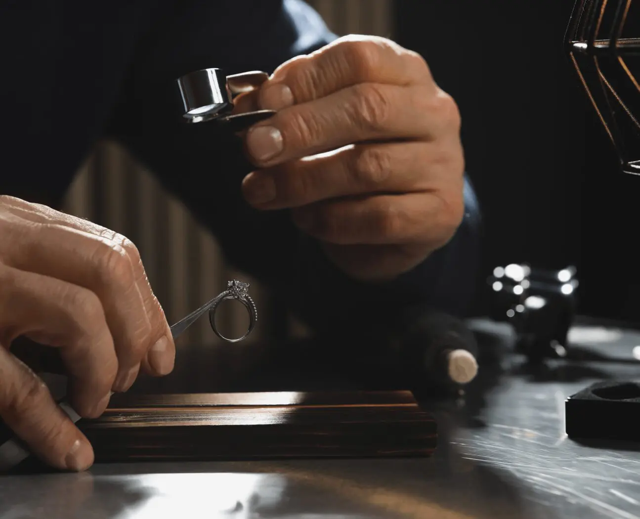 Hands inspecting a diamond ring with a magnifying loupe under a soft light, on a reflective surface with tools in the background.